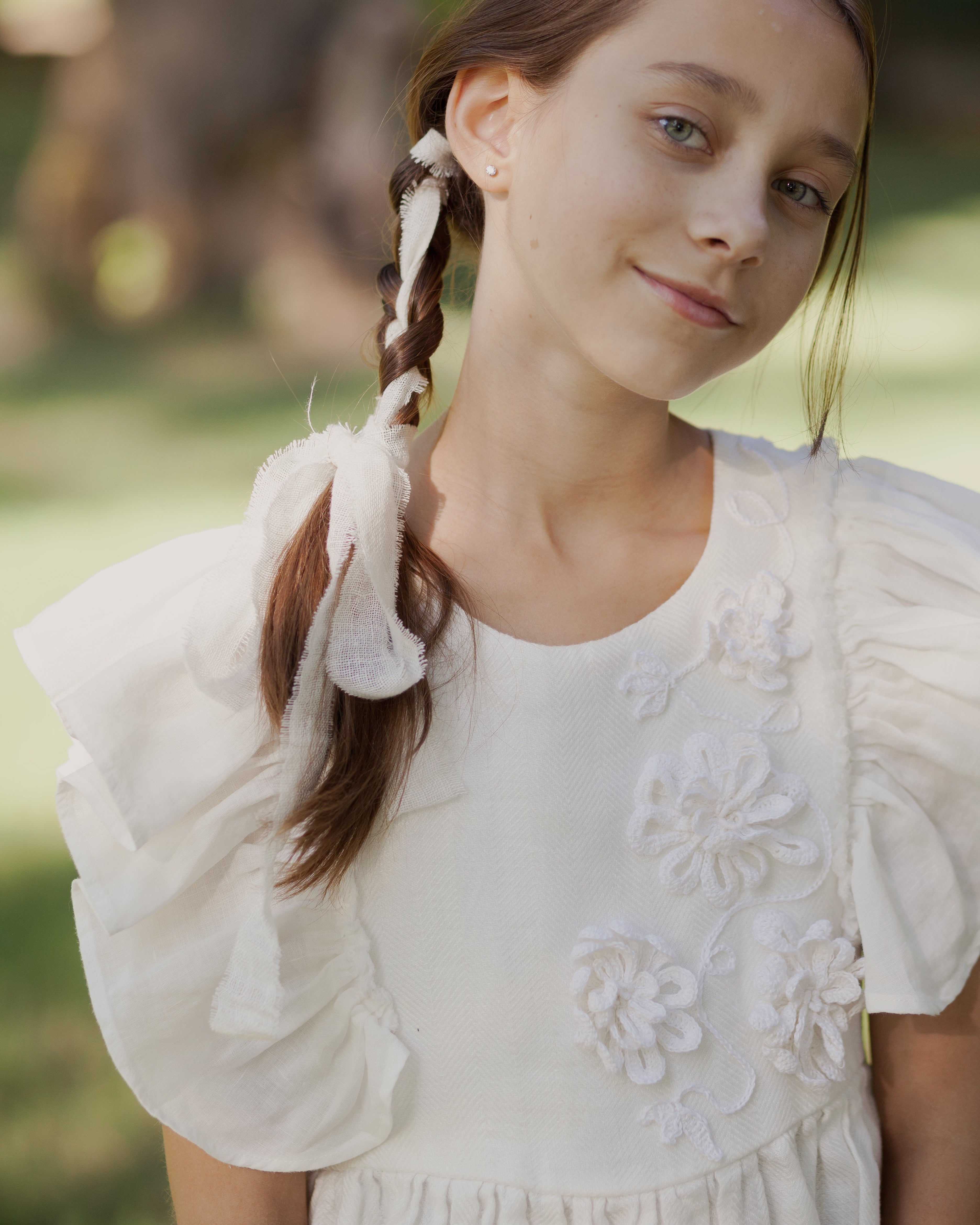 White linen dress with crochet flowers, Romantic heirloom clothing for girl, White special occasion outfit, Baptism dress for toddler, A young girl with brown hair in a side braid wearing a white linen ruffled dress with crochet floral appliques, photographed outdoors against a soft, blurred green and brown background.