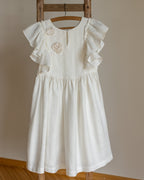 White linen dress with crochet flowers, Romantic heirloom clothing for girl, White special occasion outfit, Baptism dress for toddler, An white linen girl's dress with ruffled cap sleeves and crochet floral details on the chest, hanging on a wooden hanger from a rustic wooden ladder against a neutral beige wall.