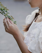 White linen dress with crochet flowers, Romantic heirloom clothing for girl, White special occasion outfit, Baptism dress for toddler, A young girl in a white linen floral dress holding a green leafy sprig, shown in a soft-focus outdoor portrait.