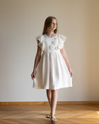White linen dress with crochet flowers, Romantic heirloom clothing for girl, White special occasion outfit, Baptism dress for toddler, A young girl wearing a white linen short-sleeve dress with ruffled sleeves and a floral crochet on the chest, standing on a wooden parquet floor against a plain beige wall.
