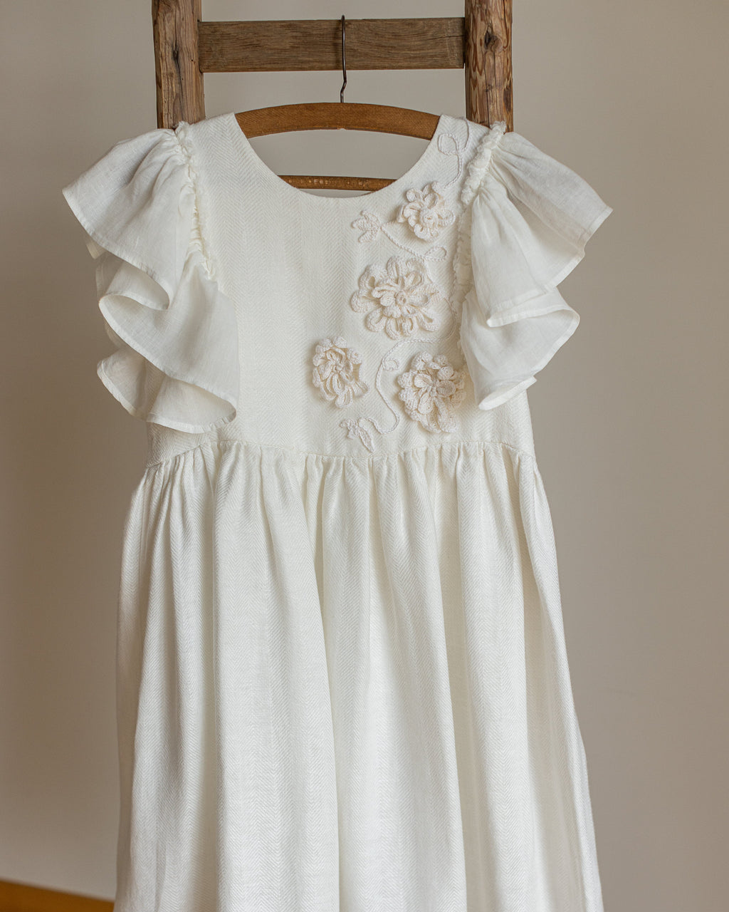 White linen dress with crochet flowers, Romantic heirloom clothing for girl, White special occasion outfit, Baptism dress for toddler, An white linen dress with ruffled cap sleeves and crochet floral applique on the chest, hanging on a wooden hanger from a rustic wooden ladder against a neutral beige wall.
