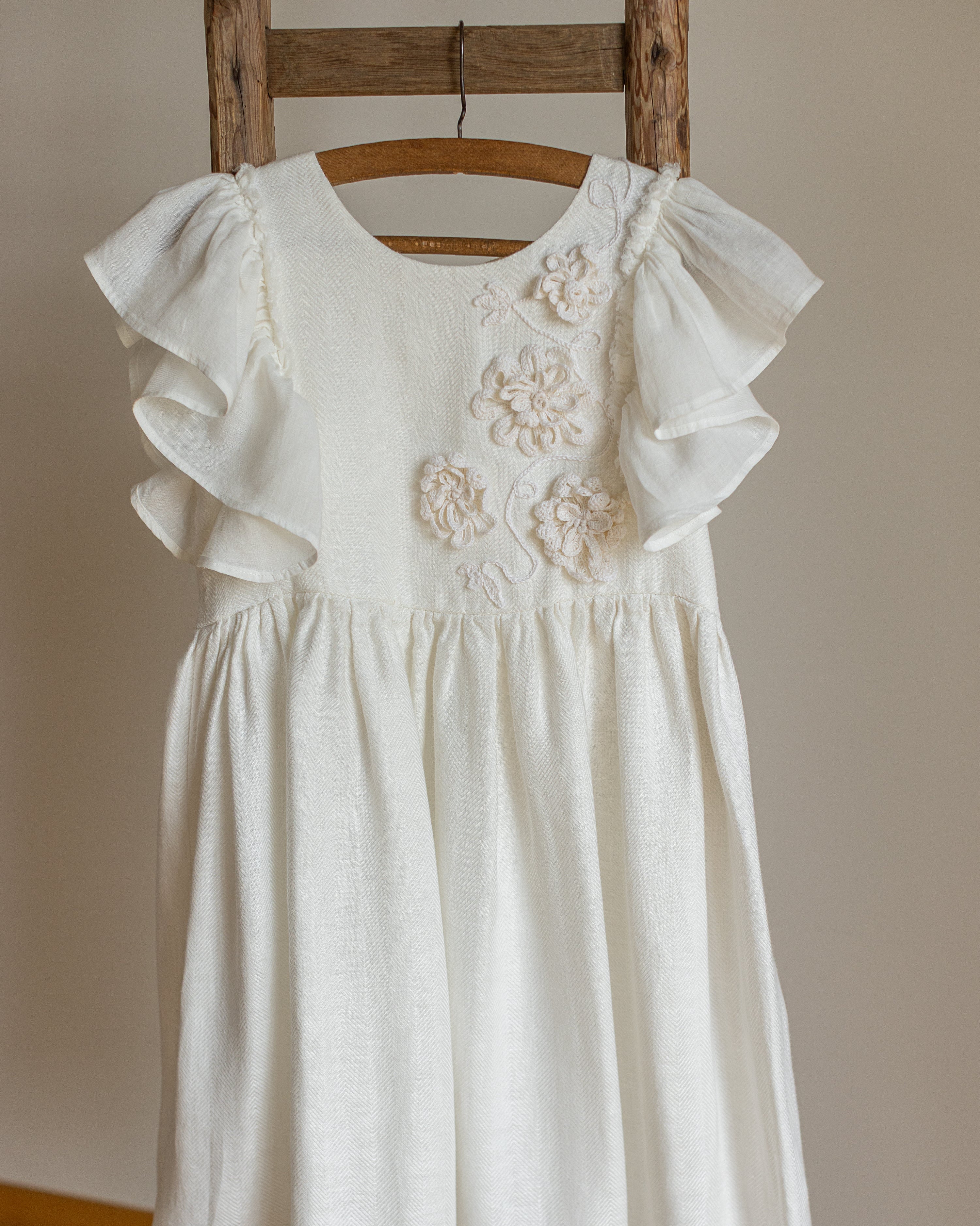 White linen dress with crochet flowers, Romantic heirloom clothing for girl, White special occasion outfit, Baptism dress for toddler, An white linen dress with ruffled cap sleeves and crochet floral applique on the chest, hanging on a wooden hanger from a rustic wooden ladder against a neutral beige wall.