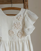 White linen dress with crochet flowers, Romantic heirloom clothing for girl, White special occasion outfit, Baptism dress for toddler, An white linen dress with ruffled sleeves and crochet floral details on the front, hanging on a wooden hanger from a rustic wooden chair against a neutral beige wall.