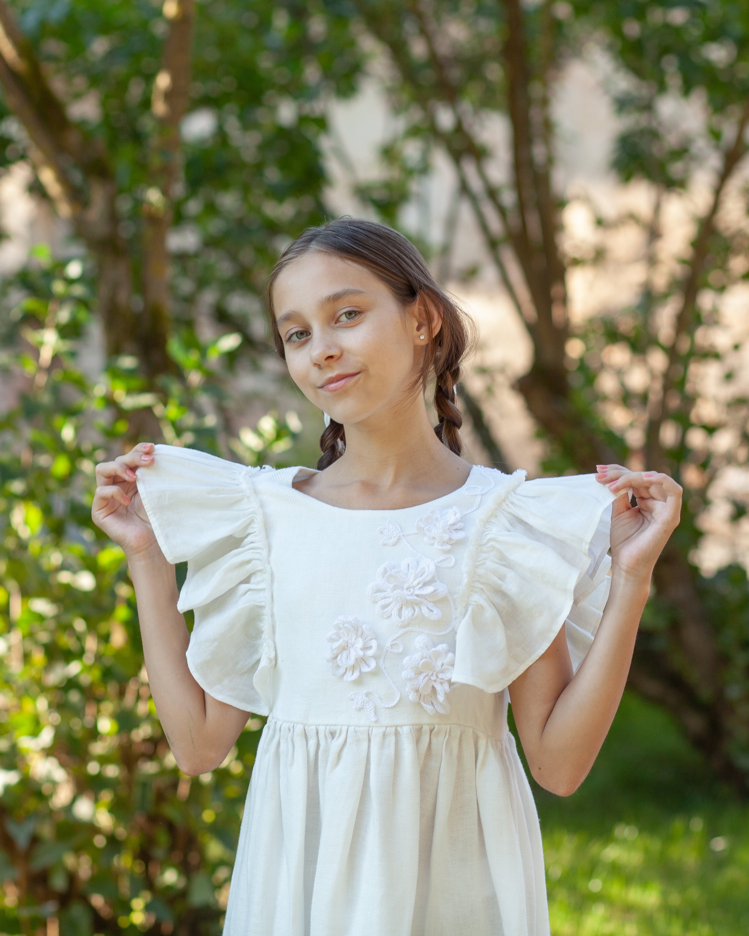 White linen dress with crochet flowers, Romantic heirloom clothing for girl, White special occasion outfit, Baptism dress for toddler, A young girl with braided hair stands outdoors in a garden, holding up a white linen dress with ruffled sleeves and floral crochet flower on the chest.