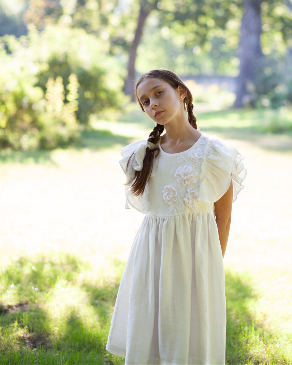White linen dress with crochet flowers, Romantic heirloom clothing for girl, White special occasion outfit, Baptism dress for toddler, A young girl with braided hair wearing a white linen dress with crochet flowers, standing in a sunny, grassy park with trees in the background.