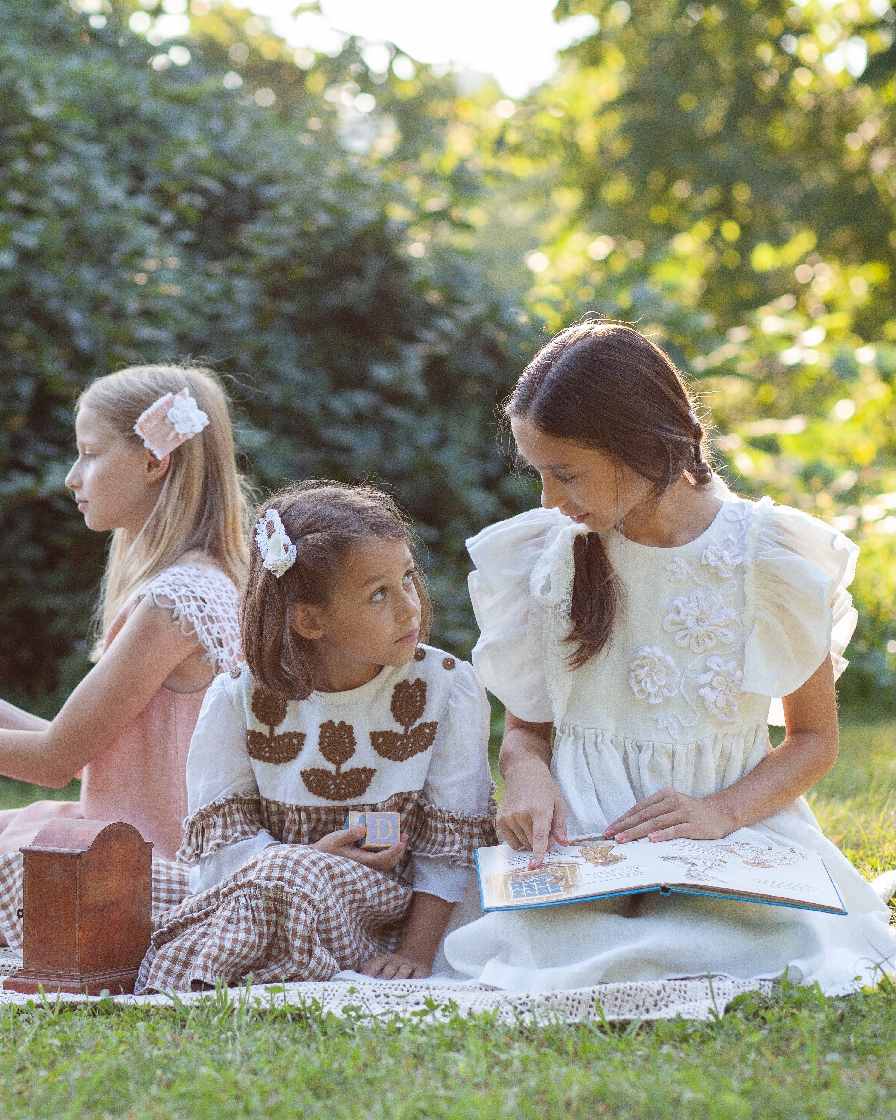 White linen dress with crochet flowers, Romantic heirloom clothing for girl, White special occasion outfit, Baptism dress for toddler, Two young girls sitting on a picnic blanket in a grassy park, reading a book together. One girl wears a white linen dress with ruffled sleeves, and the other wears a white/brown linen dress.