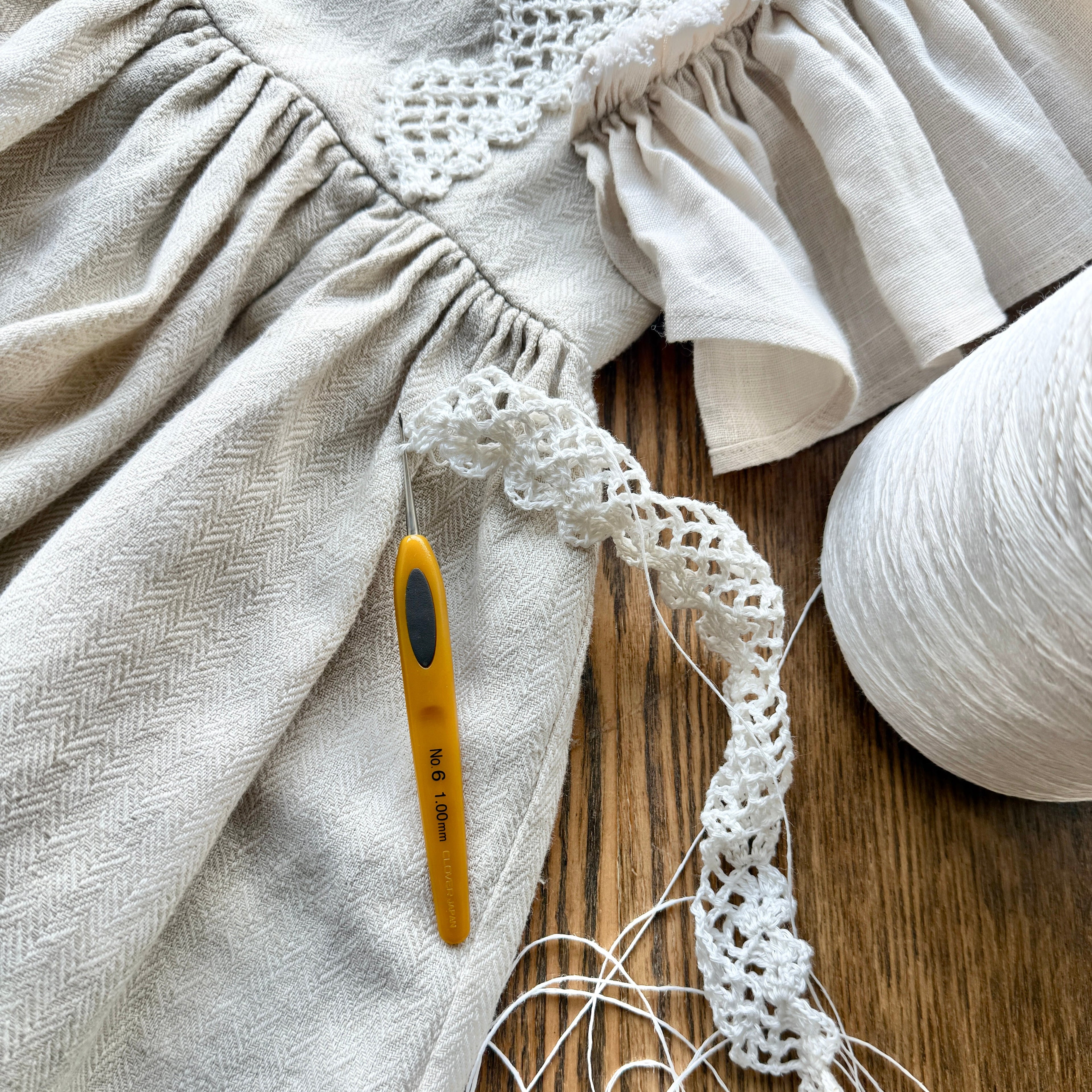 Close-up of a piece of linen dress with crochet lace trim and a crochet hook on a wooden surface.