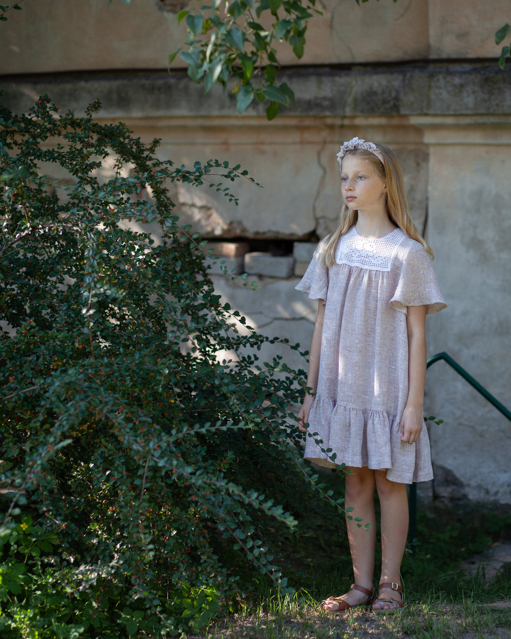 Young girl in a light linen dress standing outdoors near a stone wall and greenery