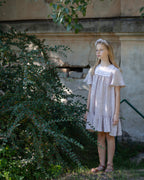 Young girl in a light linen dress standing outdoors near a stone wall and greenery