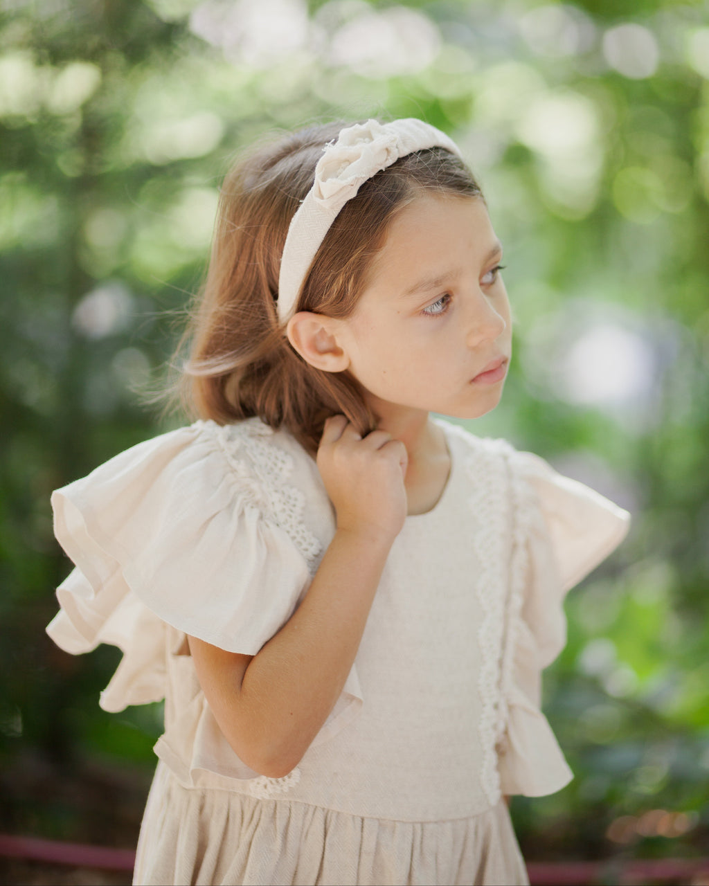 Young girl wearing a white linen dress and linen headband decorated with fabric flowers, standing outdoors with greenery in the background.
