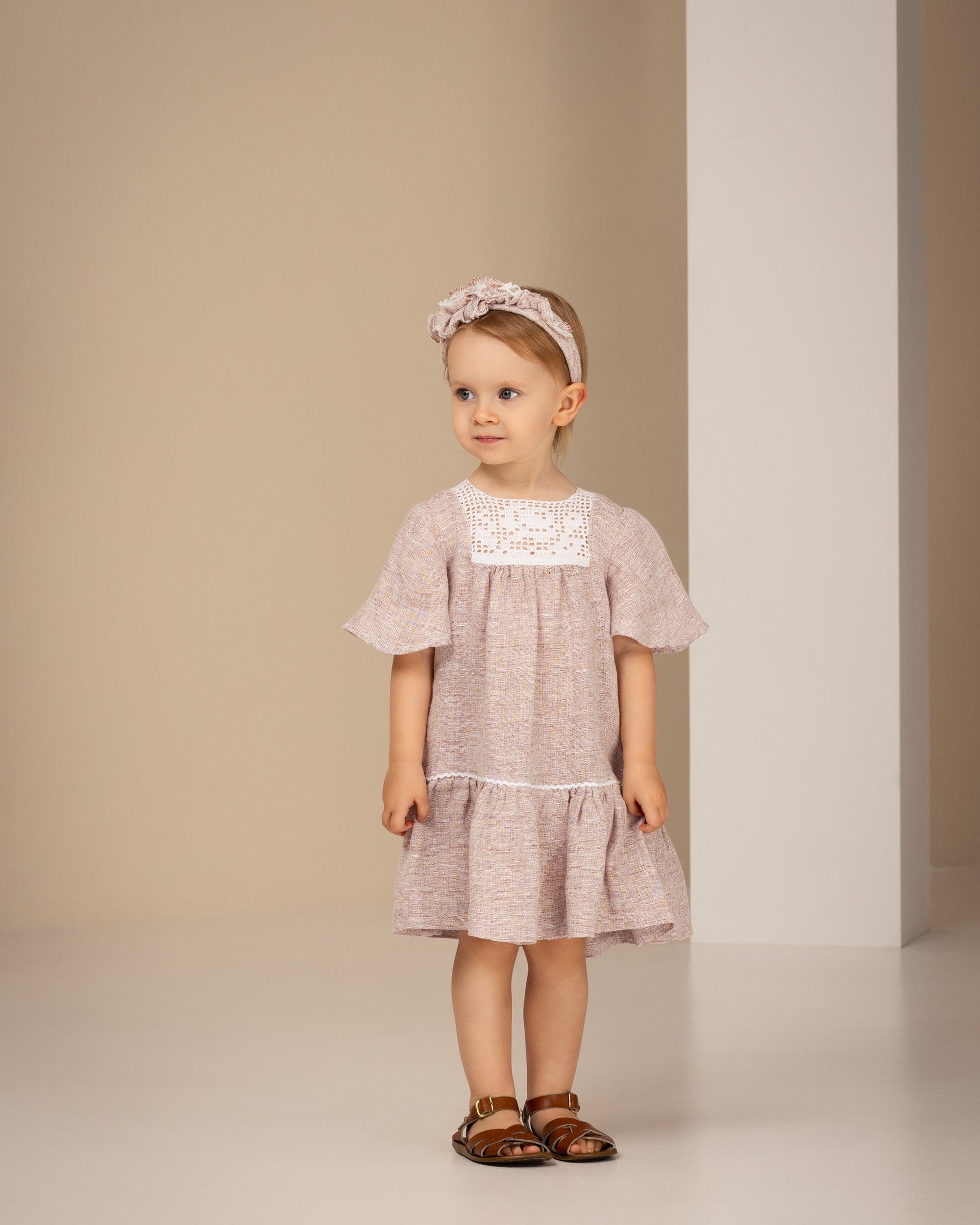Young girl wearing a light pink linen dress with lace details against a neutral background