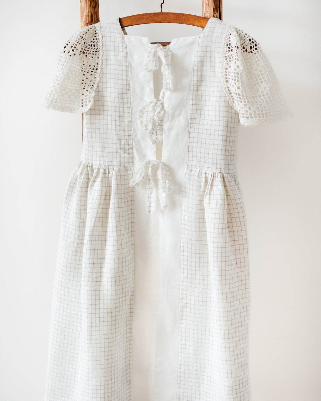 White checkered linen dress with crocheted lace sleeves on a wooden hanger against a white wall.