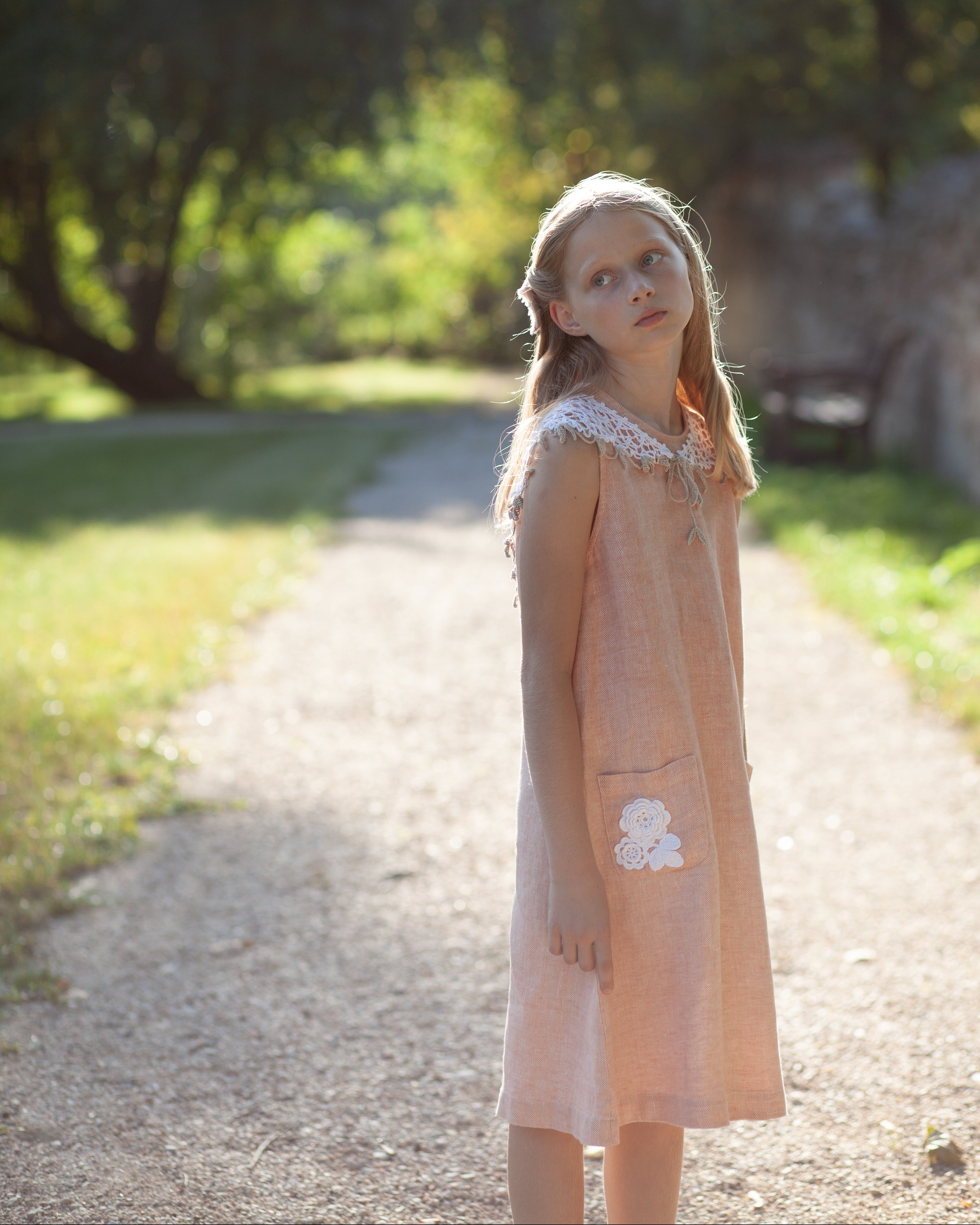 Young girl in a peach linen dress standing on a path with trees in the background