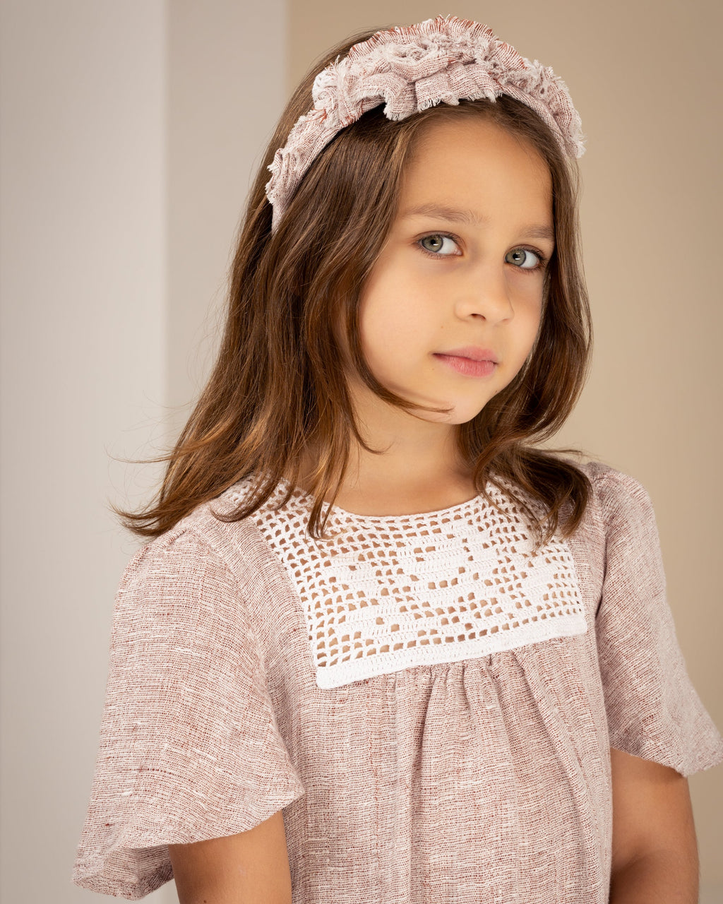 Young girl wearing a pink linen dress with crochet lace and a matching headband against a plain background
