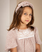 Young girl wearing a pink linen dress with crochet lace and a matching headband against a plain background