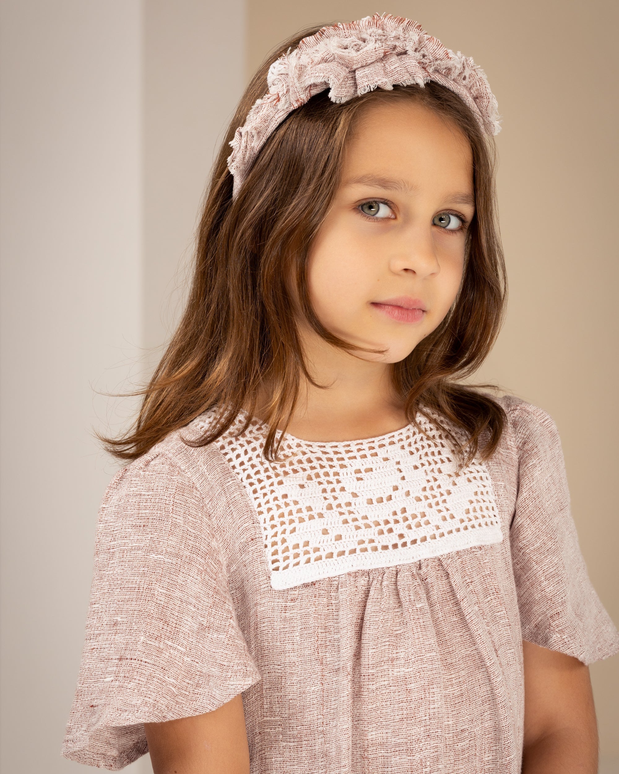 Young girl wearing a pink linen dress with crochet lace and a matching headband against a plain background