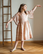 Young girl in a linen botanical dyed dress standing in a room with wooden flooring and a ladder against the wall.