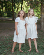 Two young girls in white linen dresses standing outdoors under a tree.