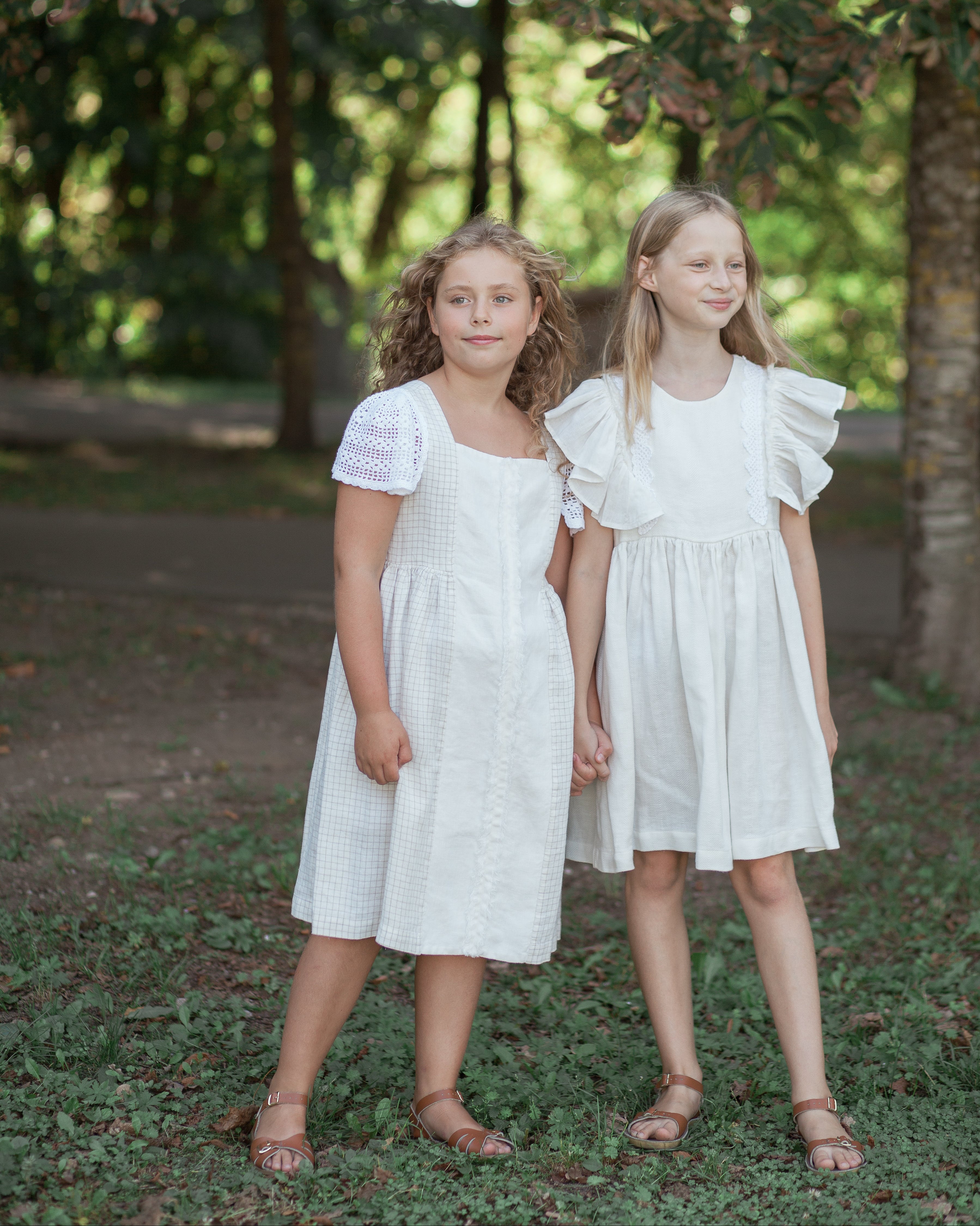 Two young girls in white linen dresses standing outdoors under a tree.