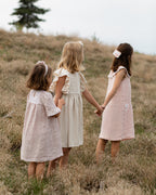 Three young girls holding hands in a field wearing matching linen dresses.