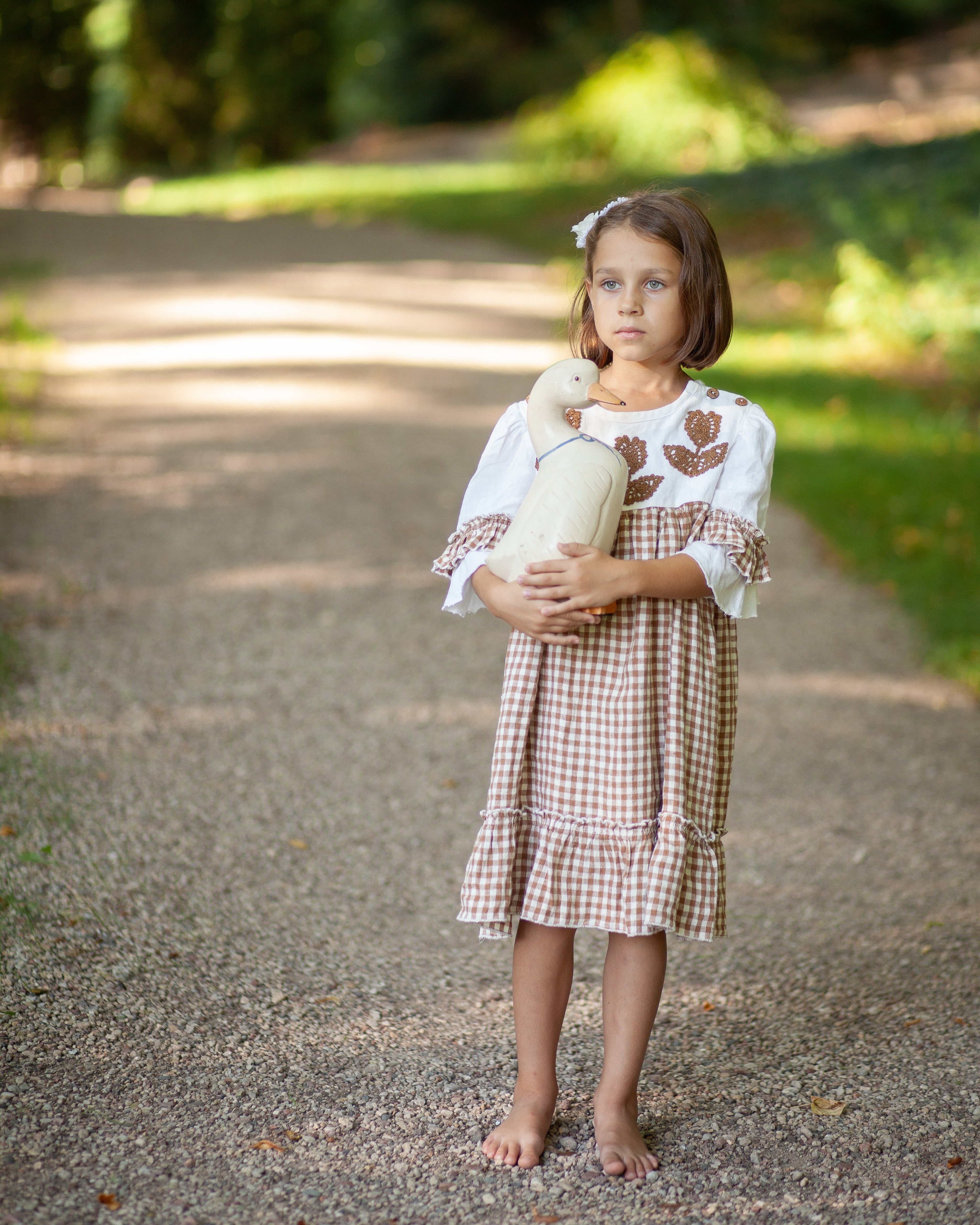 Young girl with linen dress holding a white goose on a path with greenery