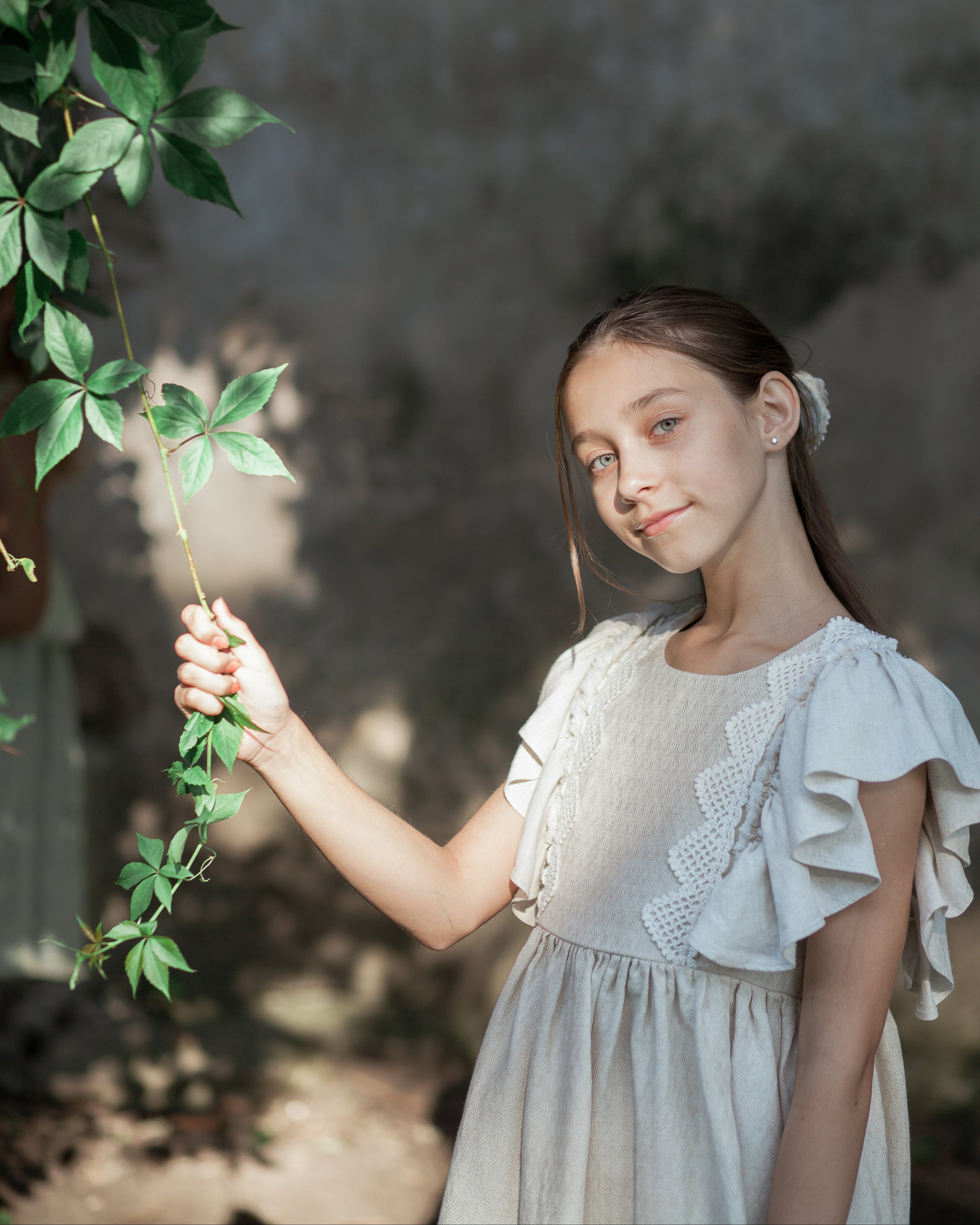 Young girl in a light sand linen dress with crochet lace holding green leaves against a blurred natural background