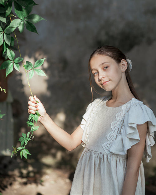 Young girl in a light sand linen dress with crochet lace holding green leaves against a blurred natural background