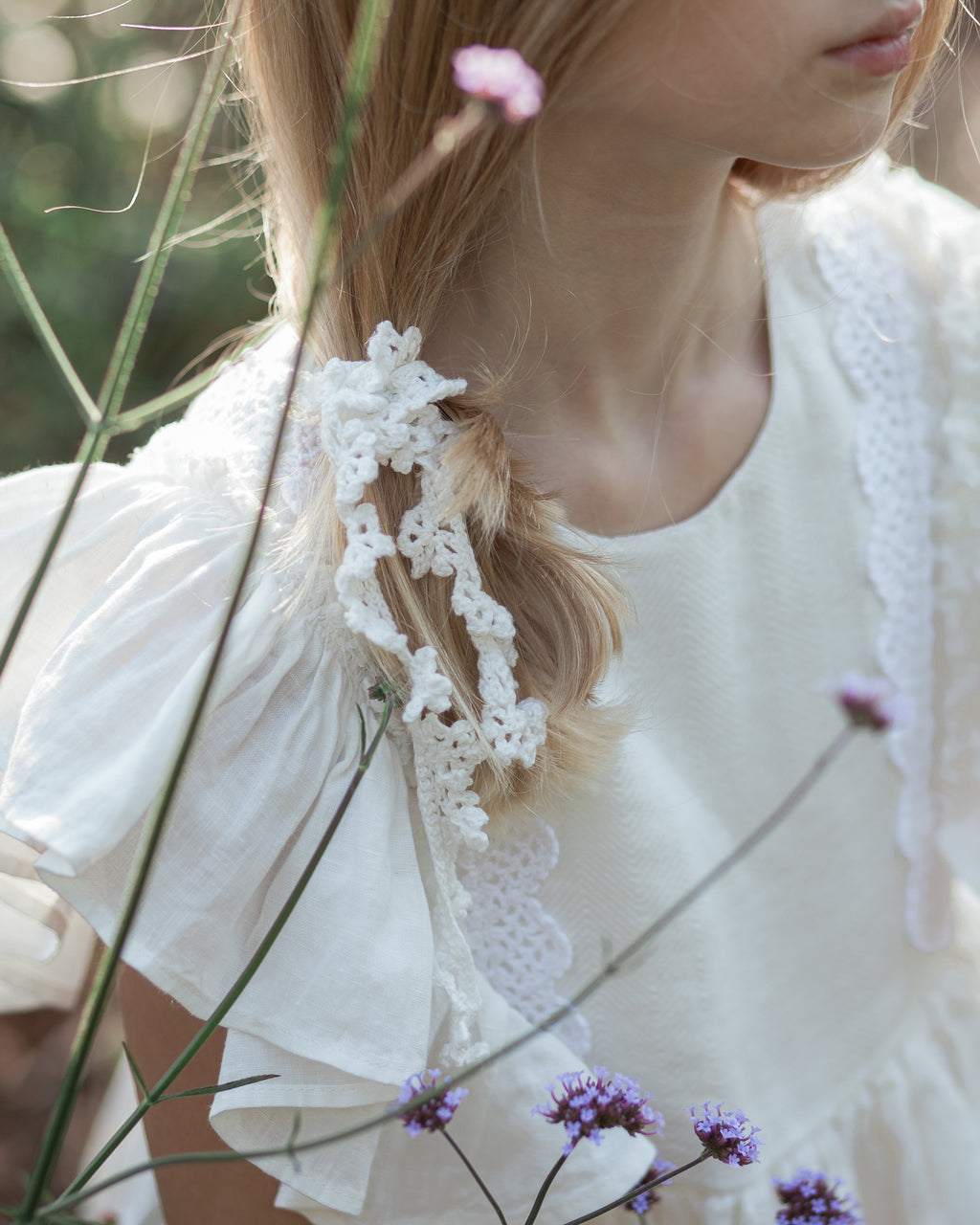 Young girl with braided hair wearing a white crochet lace headband, surrounded by flowers