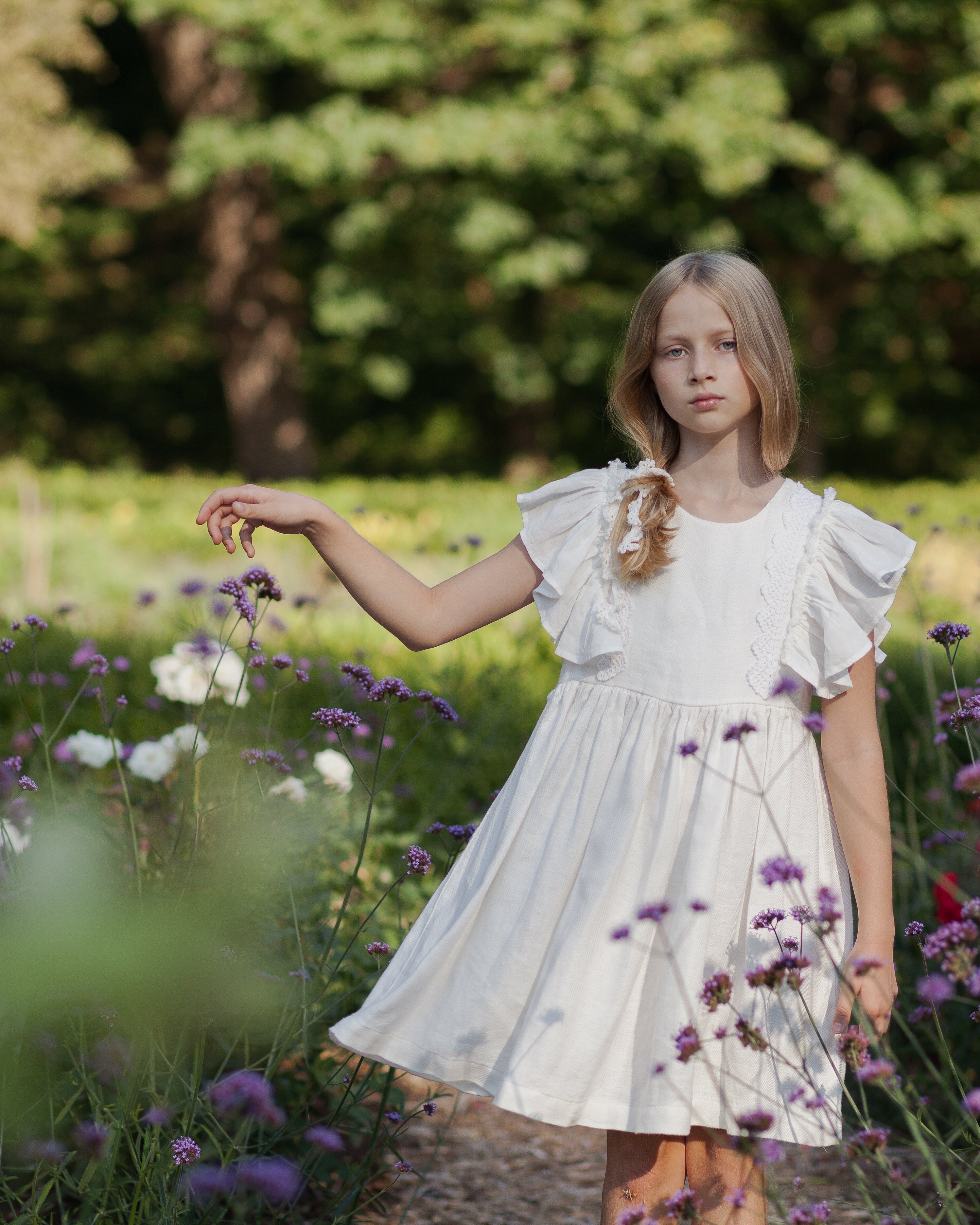 Young girl in a white linen dress with ruffled sleeves and crochet lace standing in a field of flowers with trees in the background