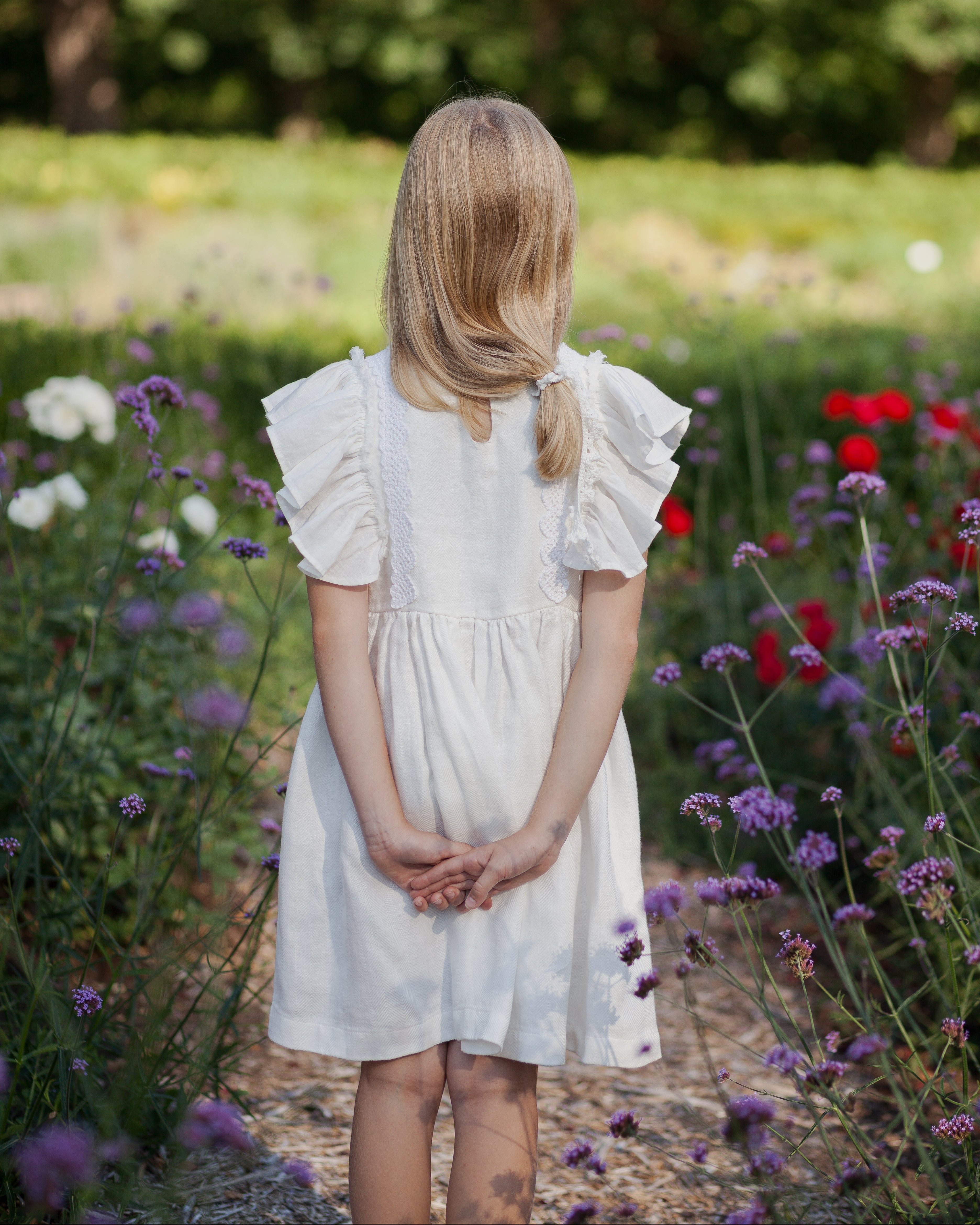 Young girl in a white linen dress with ruffled sleeves and crochet lace standing in a garden with flowers