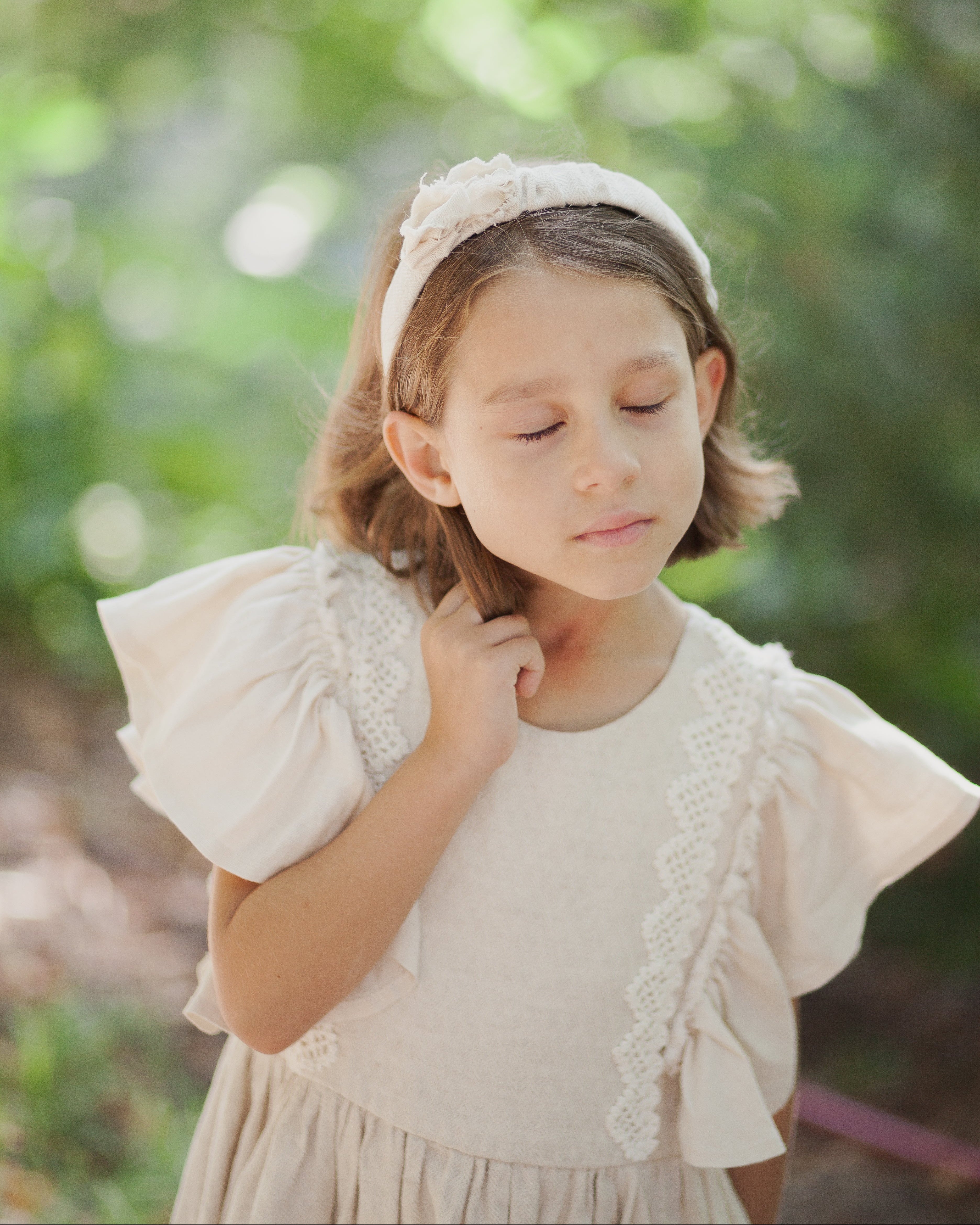Young girl wearing a light sand linen dress with ruffled sleeves and crochet lace against a blurred green background