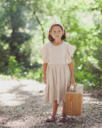 Young girl in a beige linen dress with crochet lace and headband holding a wicker suitcase in a forest setting