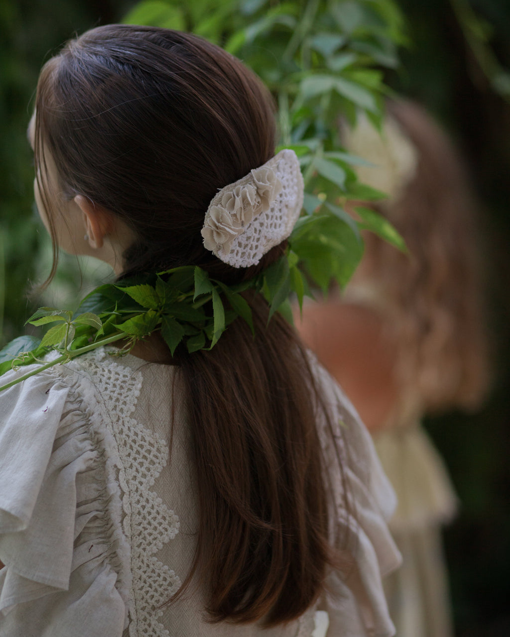 Girl with long dark hair tied back with a linen hair clip, wearing a beige linen dress with crochet lace, standing outdoors with greenery.