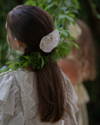 Girl with long dark hair tied back with a linen hair clip, wearing a beige linen dress with crochet lace, standing outdoors with greenery.