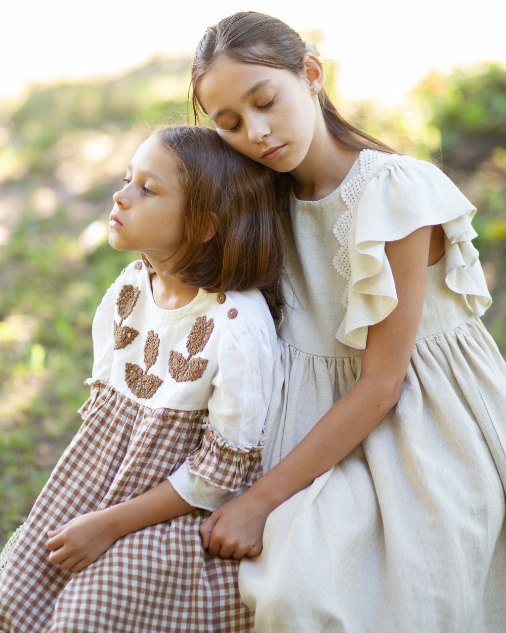 Two young girls in linen dresses sitting together outdoors with a blurred natural background