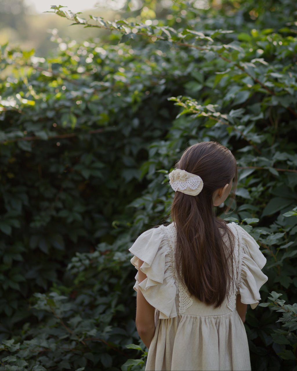 Young girl in a beige linen dress standing in front of green foliage