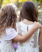 Two young girls in white linen dresses with crocheted details standing back-to-back in a field of flowers.