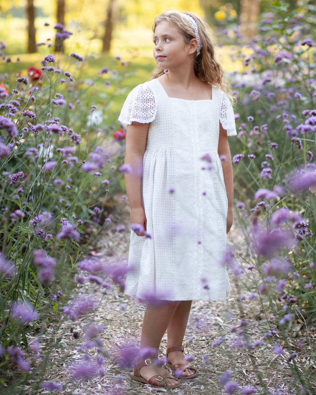 Young girl in a white linen dress with crocheted details standing in a field of purple flowers.