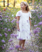 Young girl in a white linen dress with crocheted details standing in a field of purple flowers.