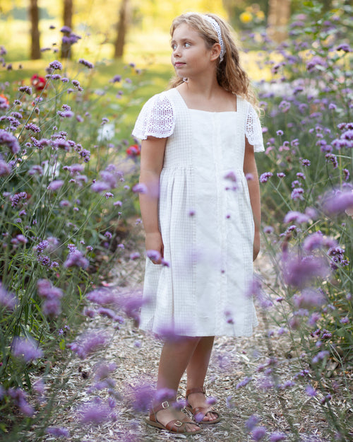 Young girl in a white linen dress with crocheted details standing in a field of purple flowers.
