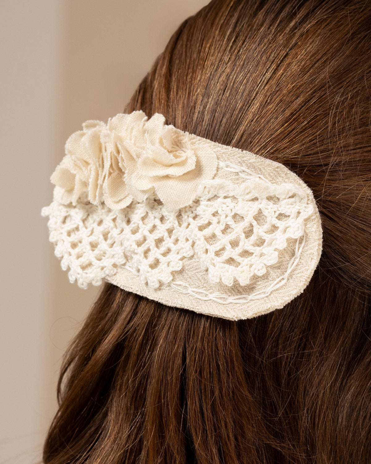 White linen hair clip with crocheted lace and fabric flowers on brown hair against a neutral background