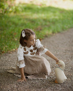 Young girl in a checkered linen dress playing with a toy goose on a path in a park.