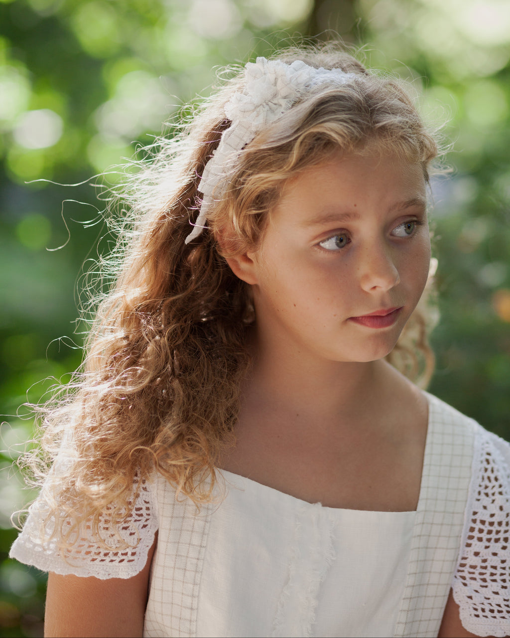 Young girl with curly hair wearing a white lace headband against a blurred green background