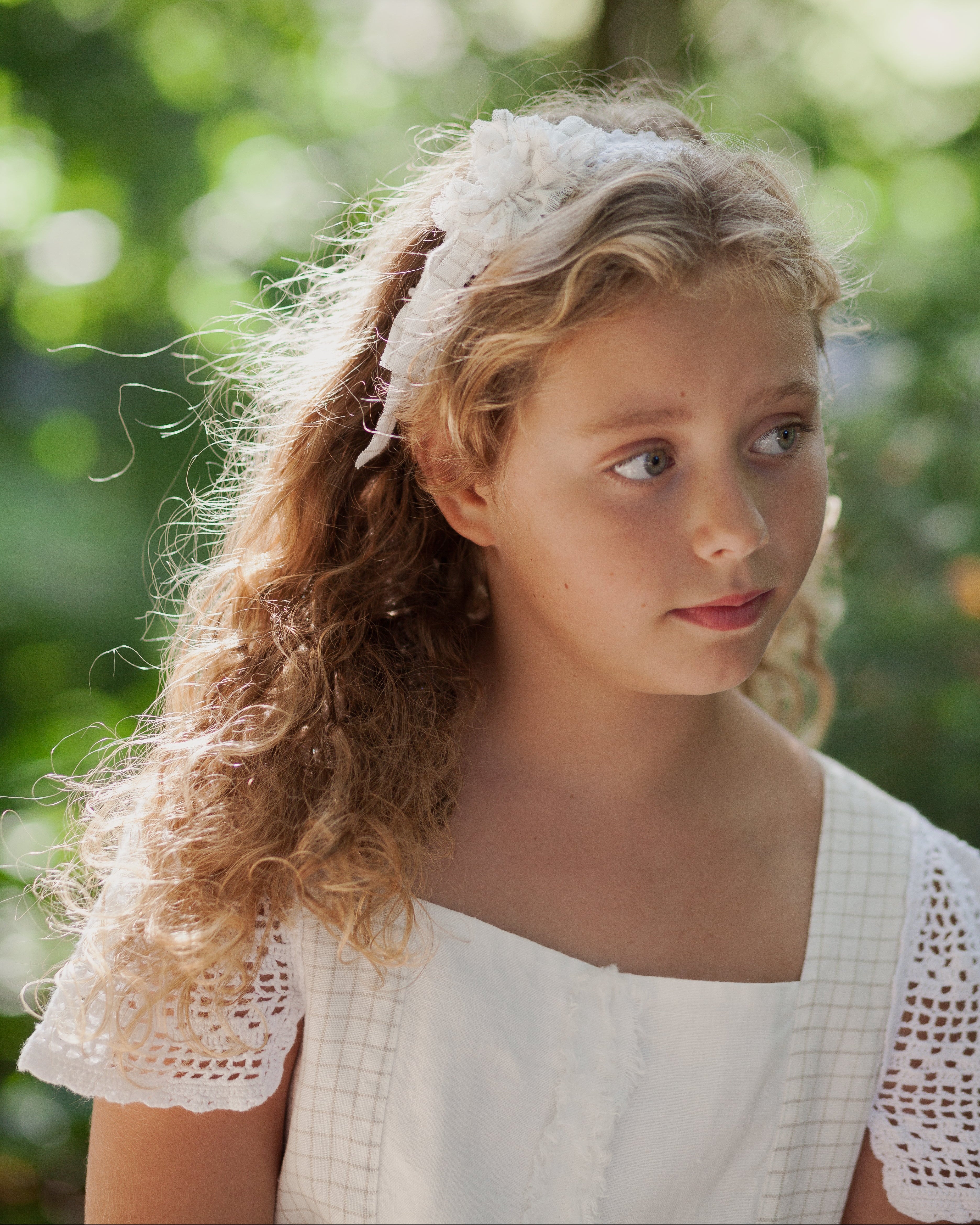 Young girl with curly hair wearing a white lace headband against a blurred green background