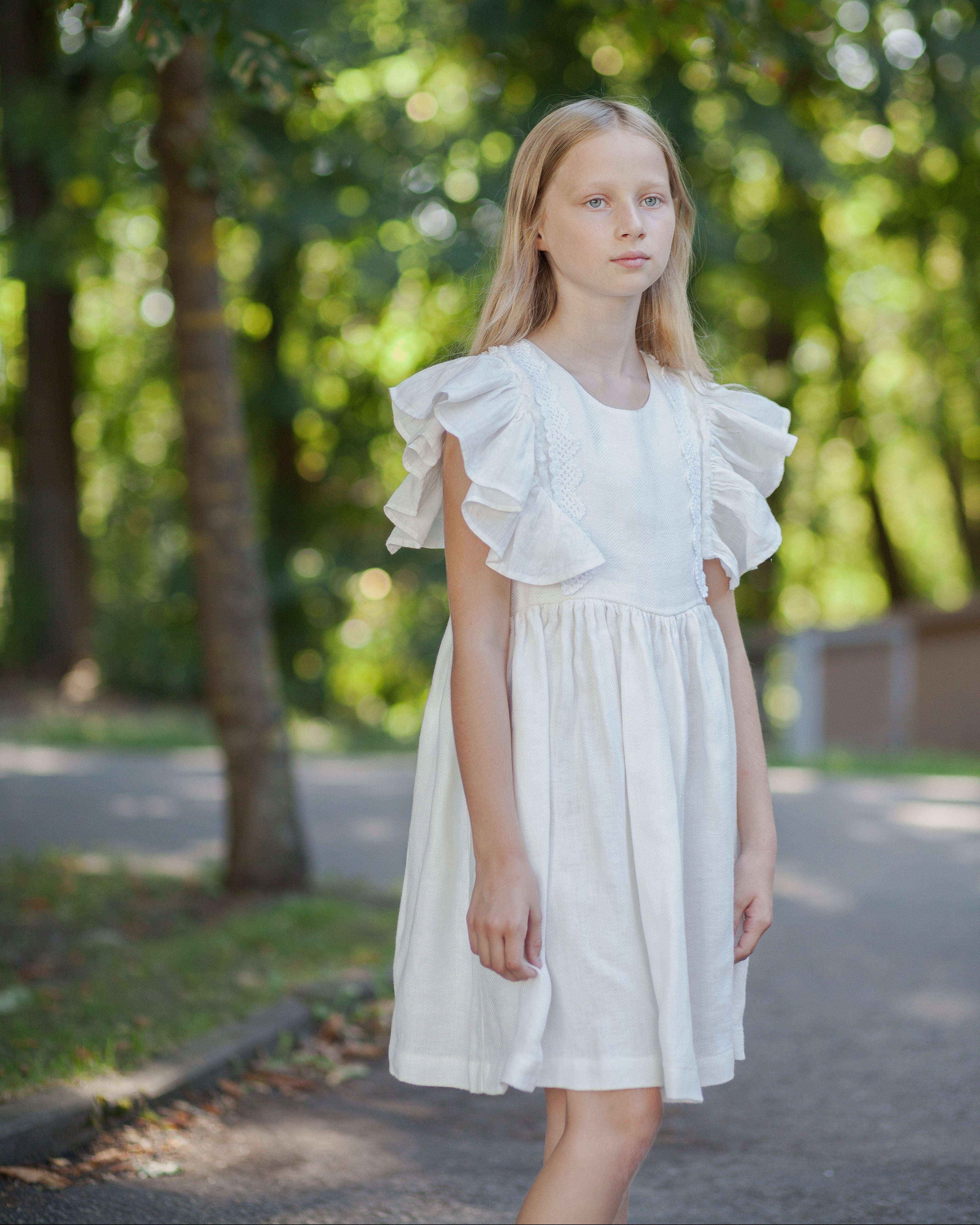 Young girl in a white linen dress with ruffled sleeves and crochet lace standing outdoors with greenery in the background