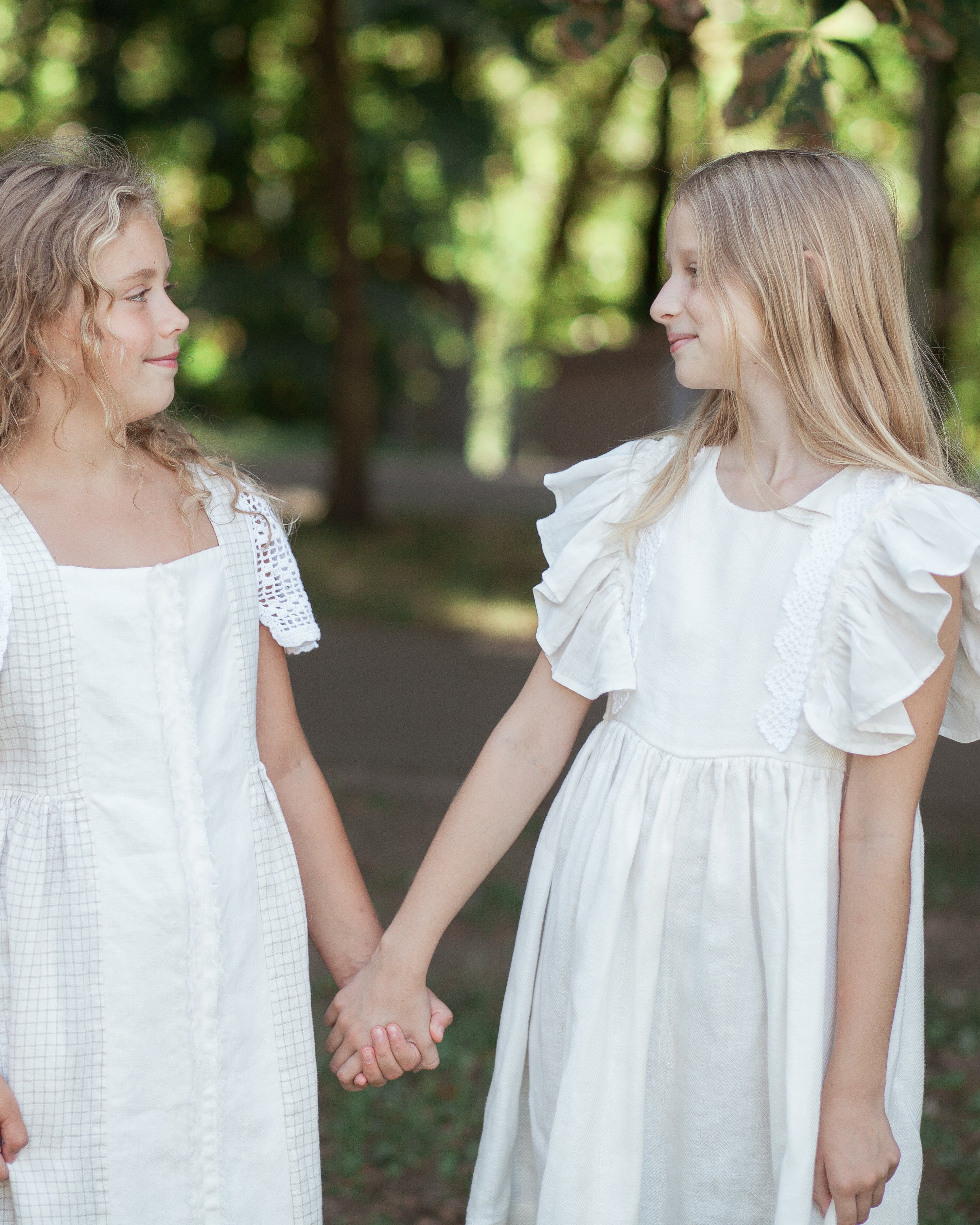 Two young girls in white linen dresses holding hands outdoors with greenery in the background