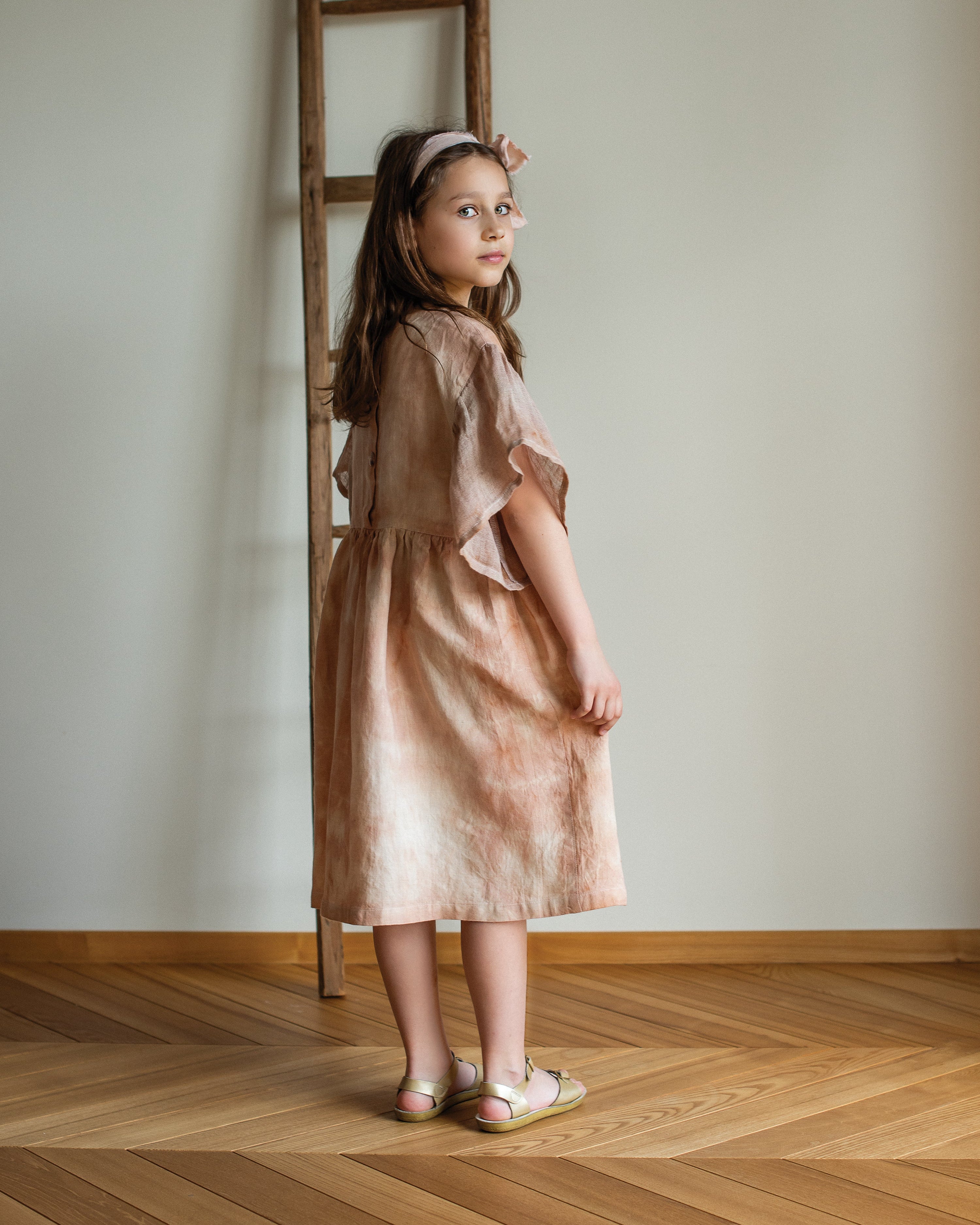 Young girl in a linen botanical dyed dress standing in a room with wooden flooring and a ladder against the wall.