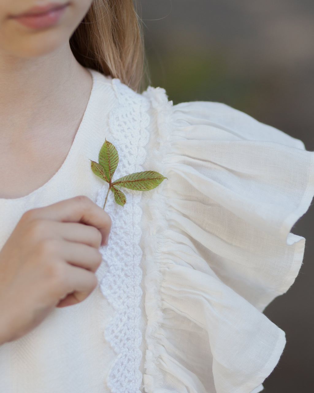 Young girl wearing a white linen dress with ruffled sleeves and crtochet details holding a small green leaf.